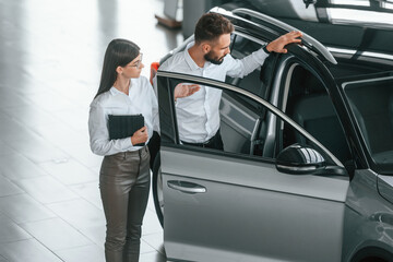 Opening the door. Man with woman in white clothes are in the car dealership together