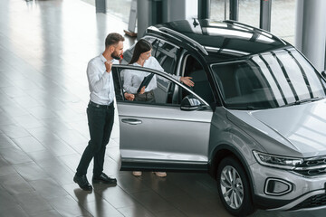 Door is opened. Man with woman in white clothes are in the car dealership together