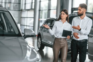 Front view. Man with woman in white clothes are in the car dealership together