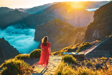 Beautiful woman in red dress walking barefoot on a very scenic hiking trail in the evening sun on Pico do Ariero. Verade do Pico Ruivo, Madeira Island, Portugal, Europe. - obrazy, fototapety, plakaty