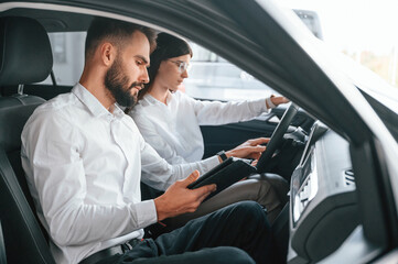 Side view. Sitting inside of automobile. Man with woman in white clothes are in the car dealership together