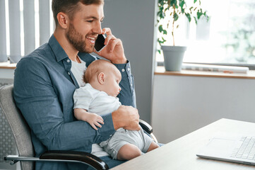 Talking by phone. Father with toddler is indoors in the office