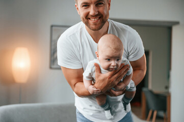 Fototapeta premium With baby in hands. Smiling, having fun. Father with toddler is at home, taking care of his son