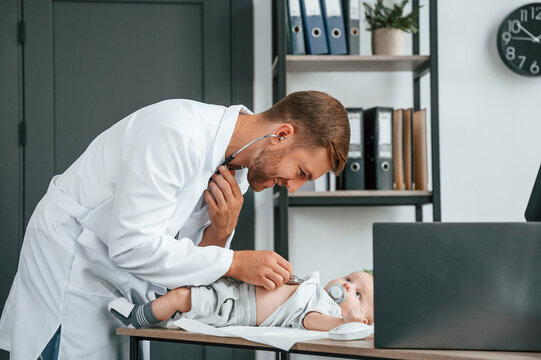 Using stethoscope. Doctor in white coat is taking care of little toddler indoors