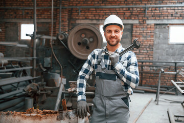 Smiling and holding wrench. Factory male worker in uniform is indoors