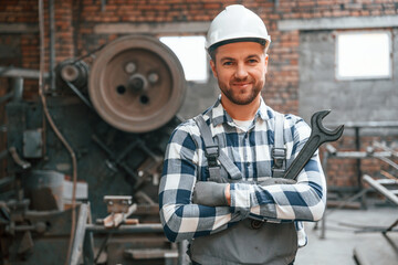 Smiling and holding wrench. Factory male worker in uniform is indoors