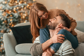 Woman is on the sofa, embracing her man by the legs. Lovely young couple are celebrating New Year at home