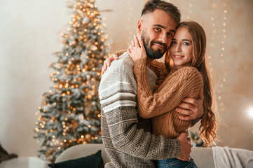 Standing against the fir with garlands on it. Lovely young couple are celebrating New Year at home