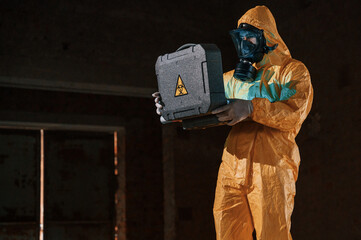 Blue light is going from hard case that is with hazard sign. Man dressed in chemical protection suit in the ruins of the post apocalyptic building