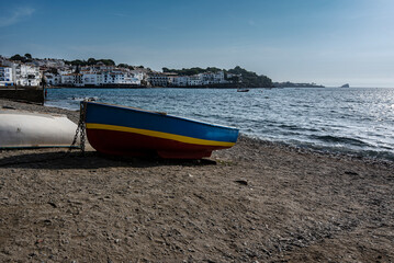 Blue, yellow, red wooden fisherman boat drying on the Cadagués pebbly beach with tha harbor and white colored houses on the background. Spain 2022