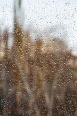 Textured surface of drops of autumn cold rain on window glass against blurred background of an empty dark city park and gray sky. Rainy weather.