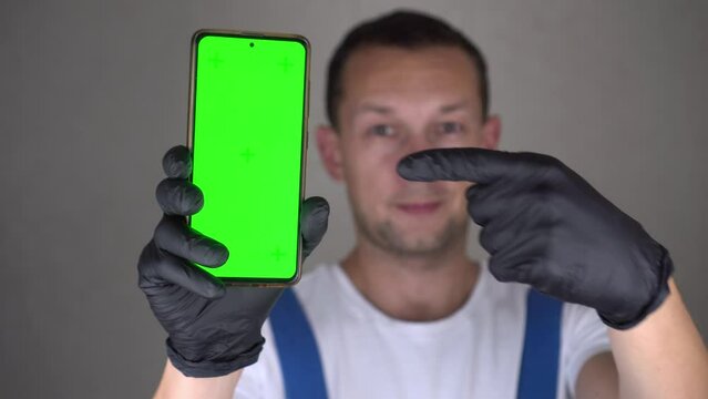 Caucasian man in blue jumpsuit holds mobile phone with green screen. Building, repair renovation of house or apartment. Person with chroma key smartphone. Professional shows app. Points with finger