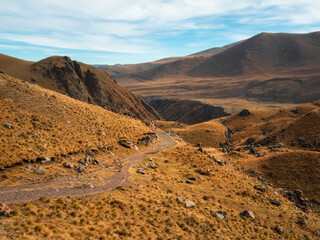 Bright sunny autumn landscape with sunlit gold valley and winding trail on mountainside under dramatic sky. Awesome alpine scenery with beautiful Caucasus mountains in golden sunshine.