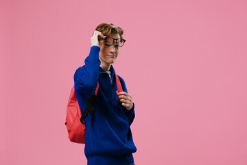 horizontal photo of a guy in a blue sweater, adjusting his glasses, with a backpack on his back. Studio photo on a uniform pink background with empty space for an advertising mockup