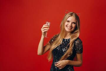 Happy blonde woman in shiny fashion clothes is happy, holding glass of champagne and posing on red background.