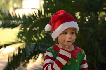 The child is waiting for a miracle, surprise, gift, holiday. A boy in a Santa Claus hat and an elf costume holds a finger to his lips: 