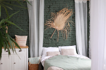 Modern boho interior of bedroom with green brick wall, bed, white wooden commode, curtains, houseplants, rattan basket, dray palm leaf and design personal accessories. Stylish home decor. Template.