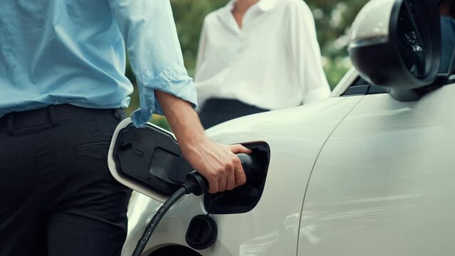 Progressive businessman and businesswoman install charger plug from charging station to electric car before driving around city center. Eco friendly rechargeable car powered by clean energy.