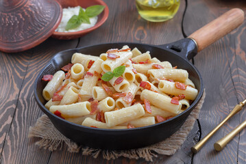 Pasta Alla Gricia.  Rigatoni pasta with bacon and pecorino cheese served in frying pan with bowl of grated cheese on background. Dark background, selective focus, horizontal.