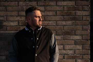 Caucasian bearded man in a bomber jacket leaned against a brick wall. 