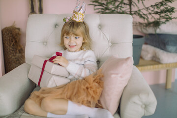 Fototapeta premium Happy pretty girl in princess crown holding Christmas present in stylish home interior decorated for winter holidays. New Year, Christmas celebration concept.