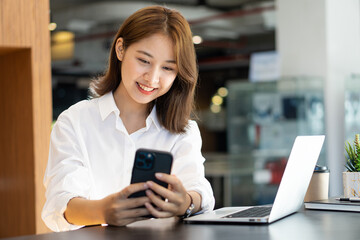 Attractive Asian woman talking on the phone and smiling while sitting at her office with laptop in the office.