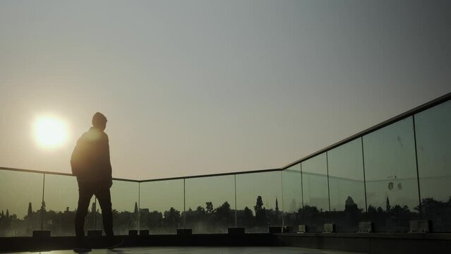 A young stylishly dressed male urban traveler with a briefcase comes to the observation deck at sunrise and admires the view. Silhouette against the background of the sun. High quality 4k footage