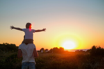 Dad carries a little son on his shoulders walking across the field against the backdrop of a sunset...