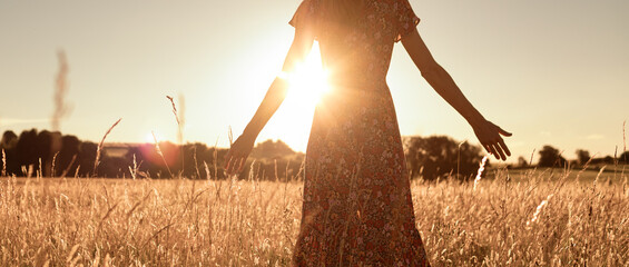 Female walking on open field at sunset softly brushing her hand over tall grass. Feeling at peace in nature concept.	 - obrazy, fototapety, plakaty