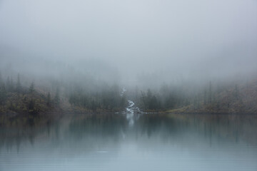 Mountain creek flows from forest hills into glacial lake in mysterious fog. Small river and coniferous trees reflected in calm alpine lake in misty morning. Tranquil landscape in fading autumn colors.