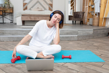 Sleepy woman doing sport and listening to music with headphones on fitness mat at home with dumbbells	