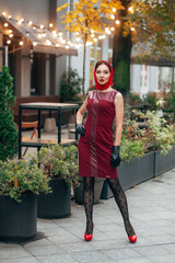 A fashionable woman in a red dress, leather gloves, tights, red shoes and a scarf, beautifully stands on the street of the evening city with illumination.