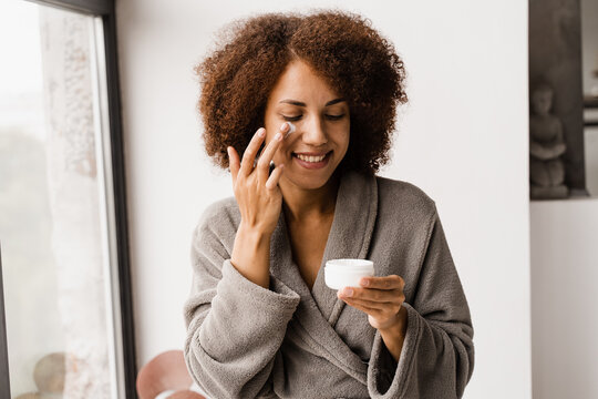 African american woman in bathrobe with facial moisturizing cream doing morning beauty routine. African girl applying face moisturizer cream to protect skin from dryness in the bath.