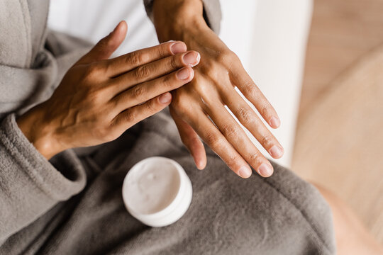 African girl applying hand moisturizer cream to protect skin from dryness in bedroom. African american woman in bathrobe with hands moisturizing cream doing morning beauty routine.