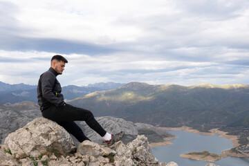 Young mountaineer dressed in dark mountain clothes on the top of the mountain is resting on top of a rock. in the background we can see a lake and the neighboring mountain range mountain range.