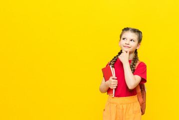A schoolgirl with a satchel looks at your advertisement for educational courses for schoolchildren. A little girl is preparing for exams on a yellow isolated background. Copy space.