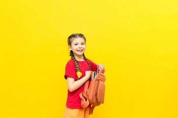 Obraz premium A schoolgirl is preparing a satchel for school. A little girl collects notebooks and textbooks for the school day on a yellow isolated background. Preparation for exams.