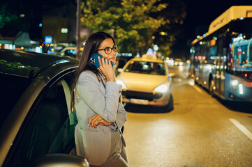 Woman calling car service on a smartphone and standing beside her car at night