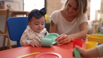 Woman and toddler learning to eat with plastic dish toy sitting on table at kindergarten