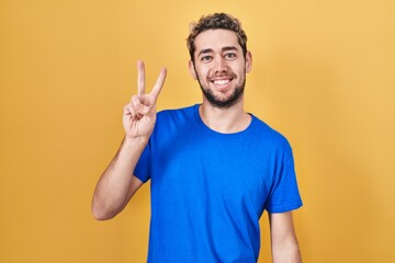 Hispanic man with beard standing over yellow background smiling with happy face winking at the...