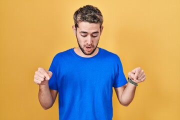 Hispanic man with beard standing over yellow background pointing down with fingers showing advertisement, surprised face and open mouth