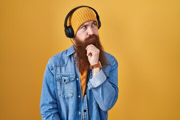 Caucasian man with long beard listening to music using headphones with hand on chin thinking about question, pensive expression. smiling with thoughtful face. doubt concept.