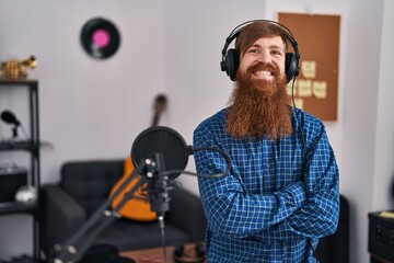 Young redhead man musician listening to music sitting with arms crossed gesture at music studio