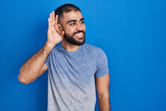 Middle east man with beard standing over blue background smiling with hand over ear listening an hearing to rumor or gossip. deafness concept.