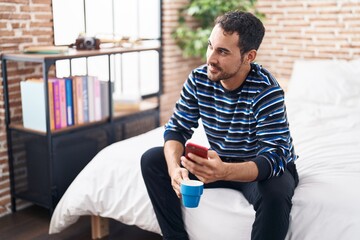 Young hispanic man using smartphone drinking coffee at bedroom