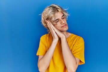 Young modern man standing over blue background sleeping tired dreaming and posing with hands together while smiling with closed eyes.