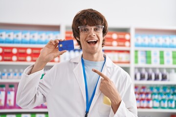 Hispanic young man working at pharmacy drugstore holding credit card smiling happy pointing with hand and finger