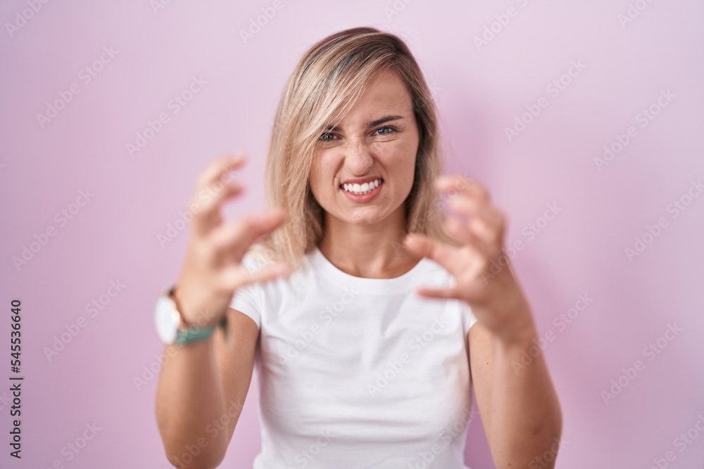 Wall mural Young blonde woman standing over pink background shouting frustrated with rage, hands trying to strangle, yelling mad