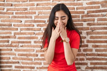 Young teenager girl standing over bricks wall laughing and embarrassed giggle covering mouth with...