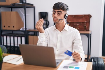 Young hispanic man working using computer laptop holding credit card doing italian gesture with hand and fingers confident expression